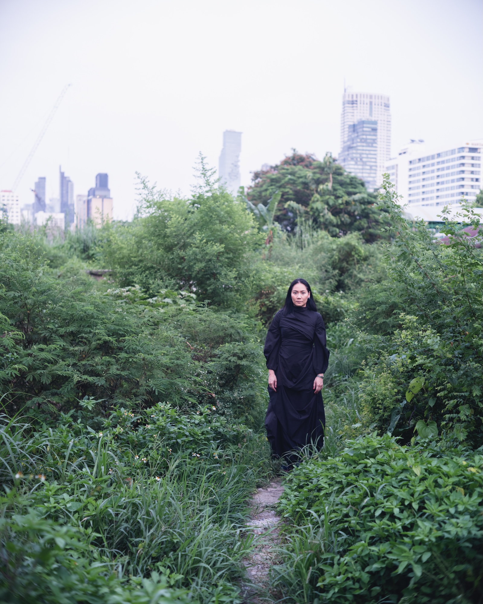 Dujdao standing in a black dress amid dense greenery, with city buildings behind her.