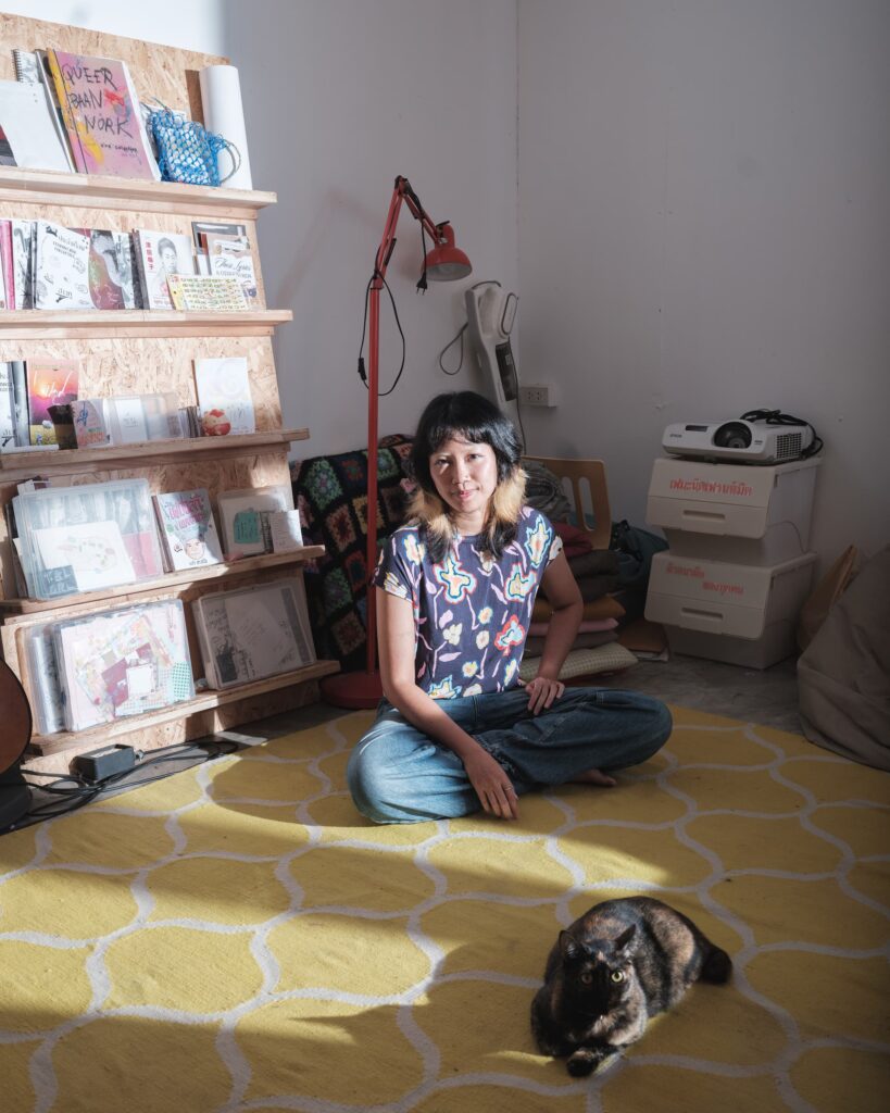 Zoom seated on a yellow rug inside Patata Studio in Khon Kaen, with her cat Phoon in the foreground and zines on shelves beside her.