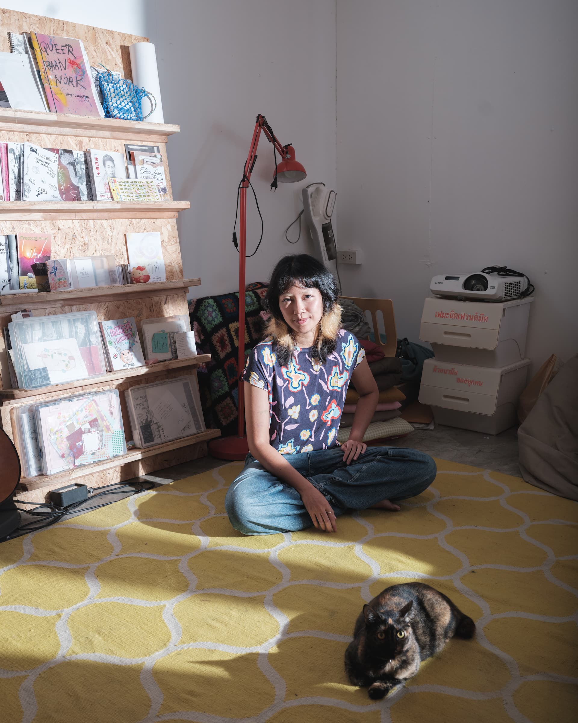 Zoom seated on a yellow rug inside Patata Studio in Khon Kaen, with her cat Phoon in the foreground and zines on shelves beside her.