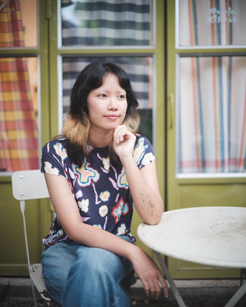 Zoom seated at a small round table outside Patata Studio in Khon Kaen, framed by green doors and striped curtains.