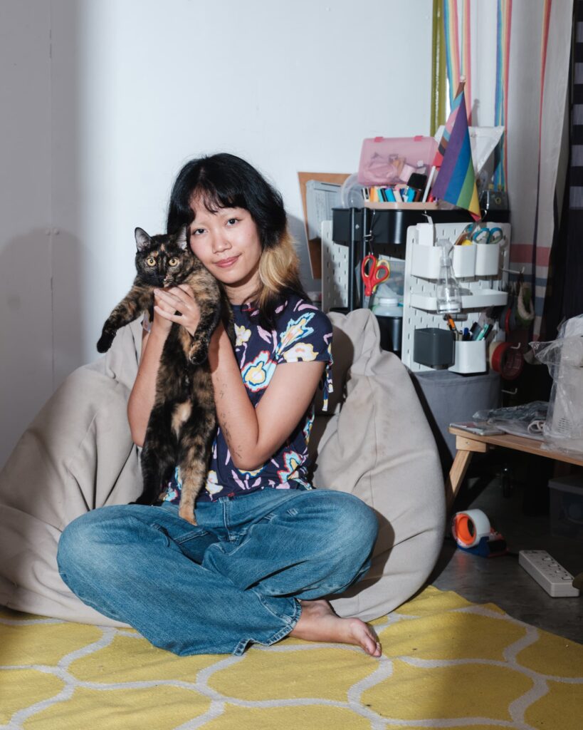 Zoom seated inside Patata Studio in Khon Kaen holding her cat Phoon, with craft tools and studio materials behind them.