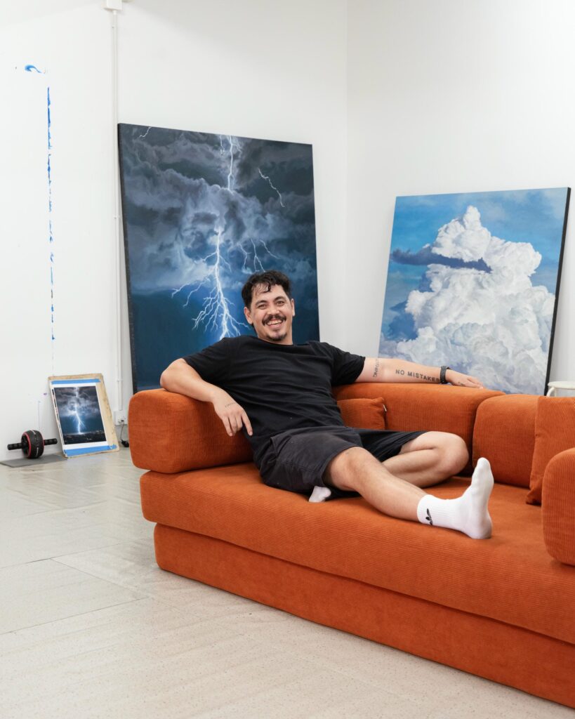 Abdul Abdullah reclining on an orange sofa in front of paintings of clouds and lightning.