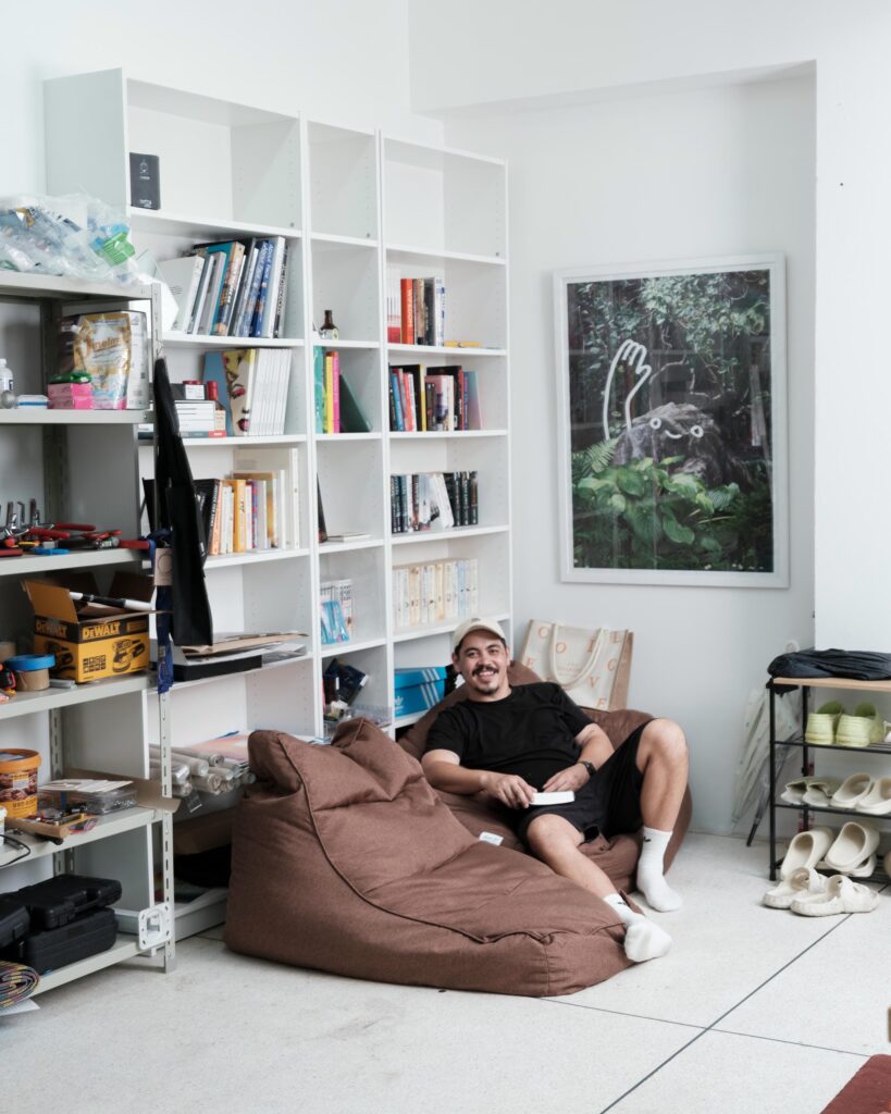 Abdul Abdullah seated on a beanbag beside shelves of books and studio materials.