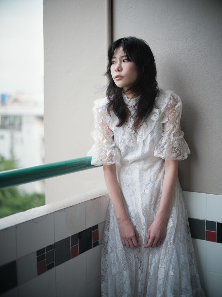 Aey Kuljira Thongkong standing beside a balcony railing in a white lace dress.
