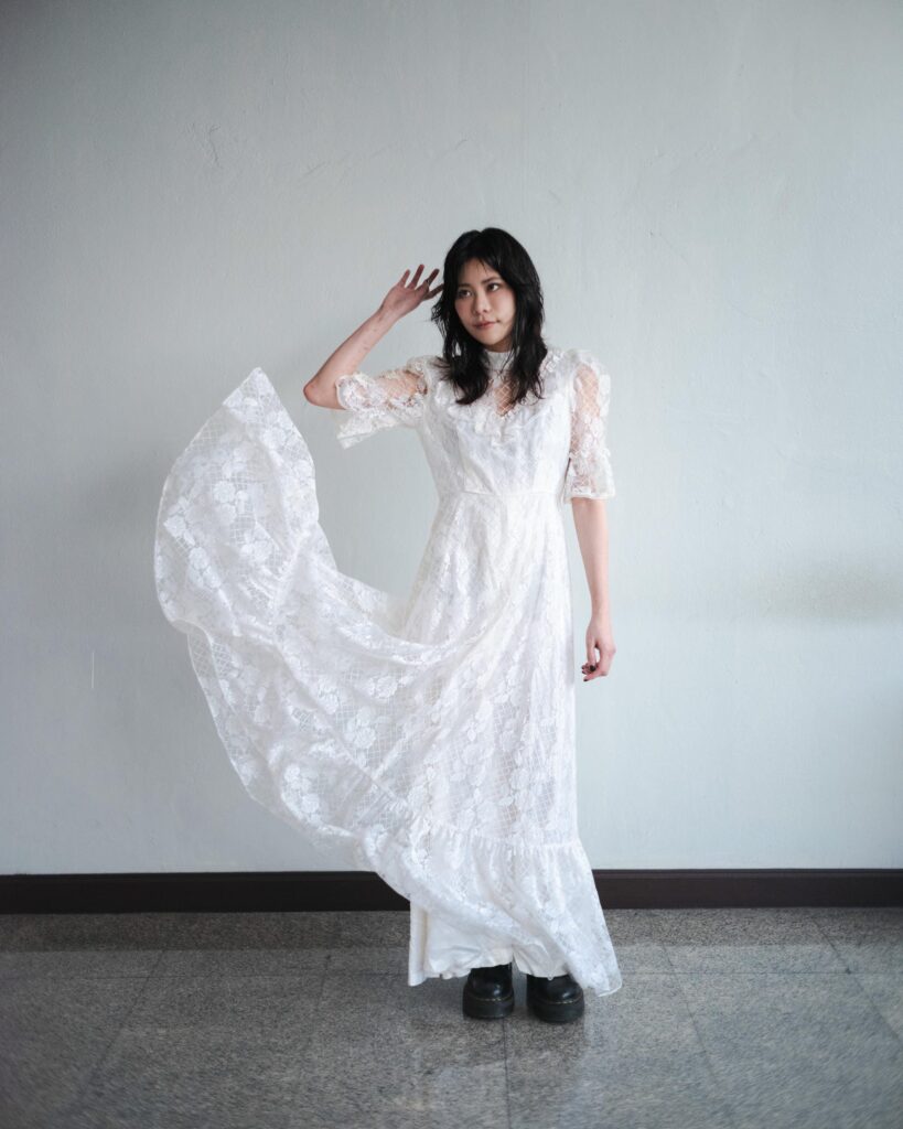 Aey Kuljira Thongkong lifting the skirt of a white lace dress against a plain wall.