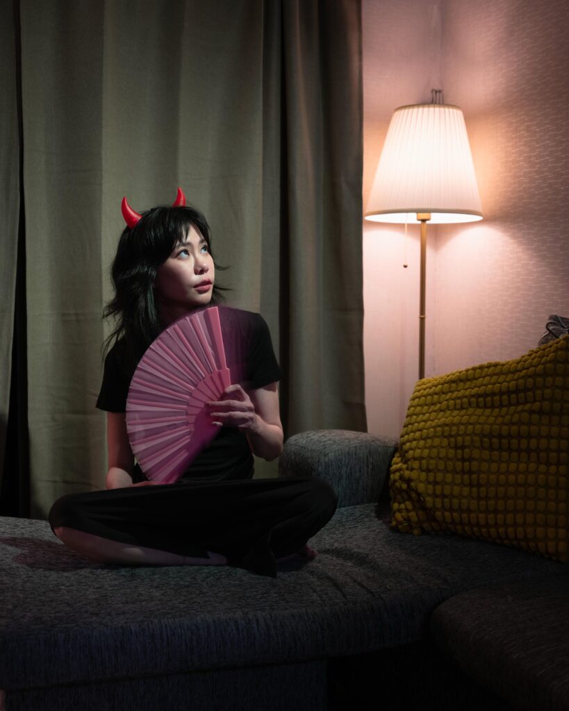 Aey Kuljira Thongkong sitting on a sofa with a pink fan beside a floor lamp.