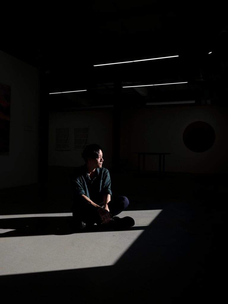 Chol Janepraphaphan seated on the gallery floor between areas of light and shadow.