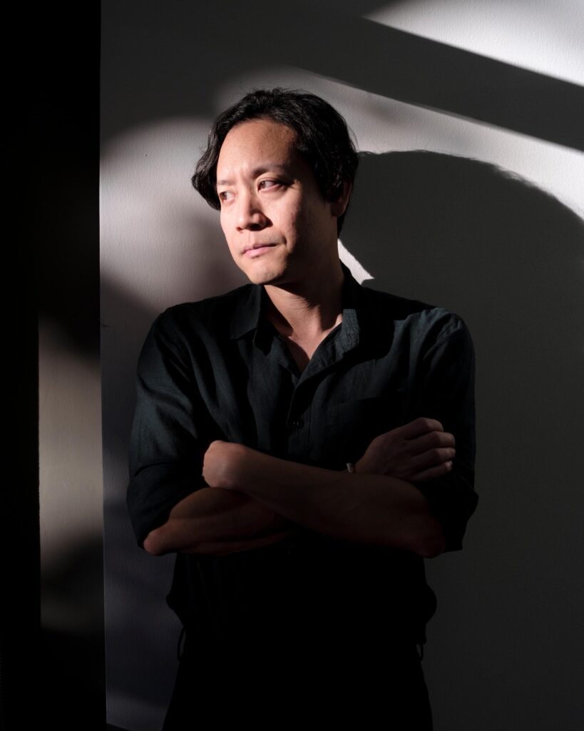 Chol Janepraphaphan standing with folded arms in sunlight against a gallery wall.