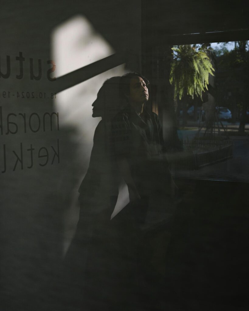 Chol Janepraphaphan's profile reflected in a gallery window with trees visible beyond.