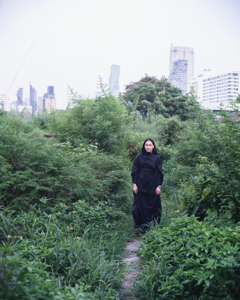 Dujdao Vadhanapakorn standing among tall plants with Bangkok buildings behind her.
