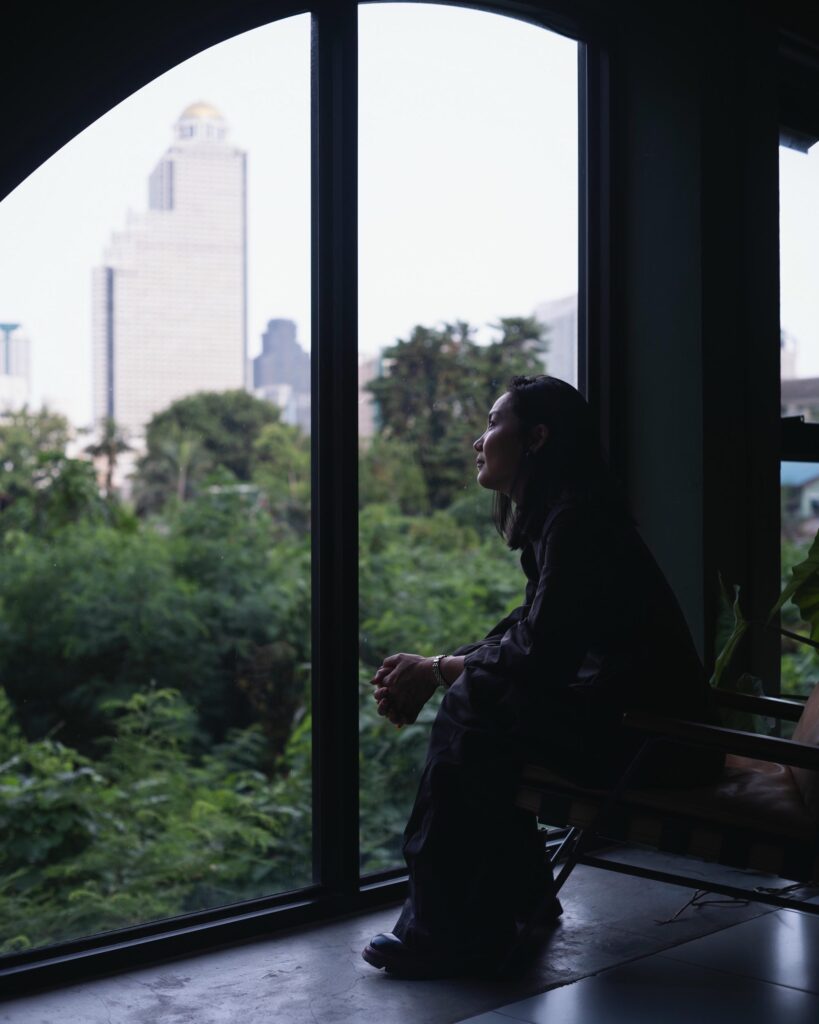 Dujdao Vadhanapakorn seated beside an arched window overlooking trees and city buildings.
