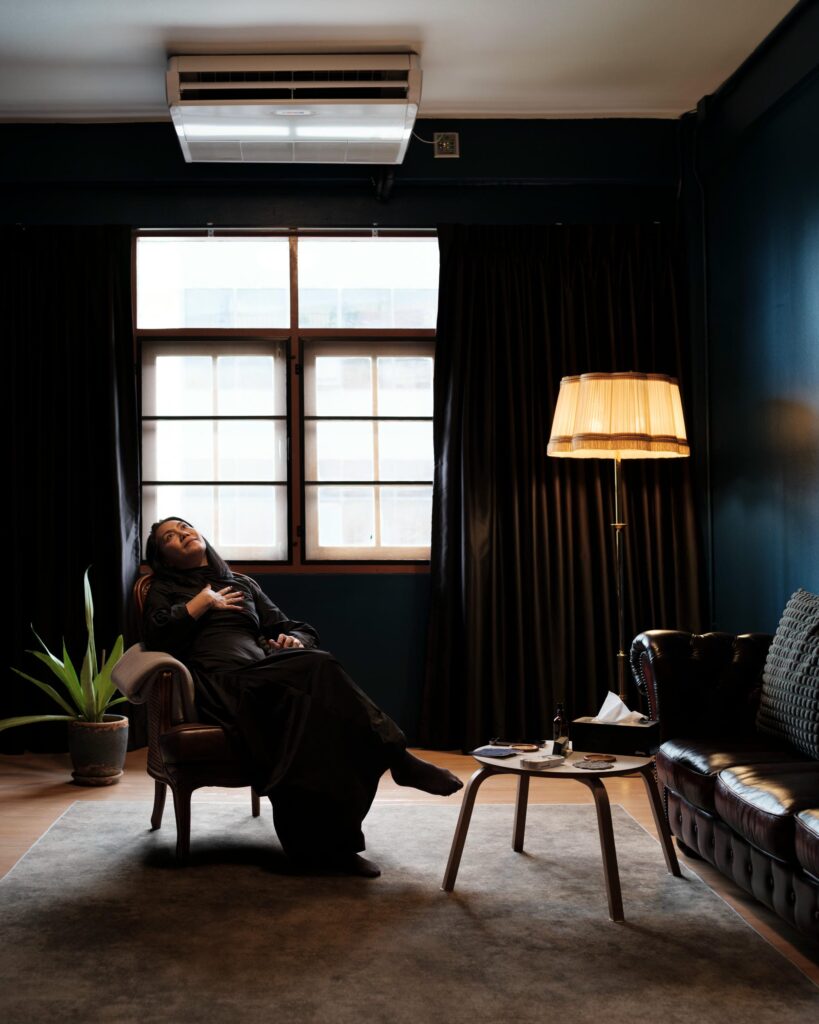Dujdao Vadhanapakorn reclining in an armchair beside a window and floor lamp.