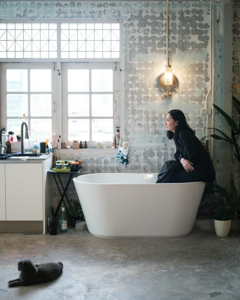 Dujdao Vadhanapakorn sitting on the edge of a freestanding bathtub beneath a window.