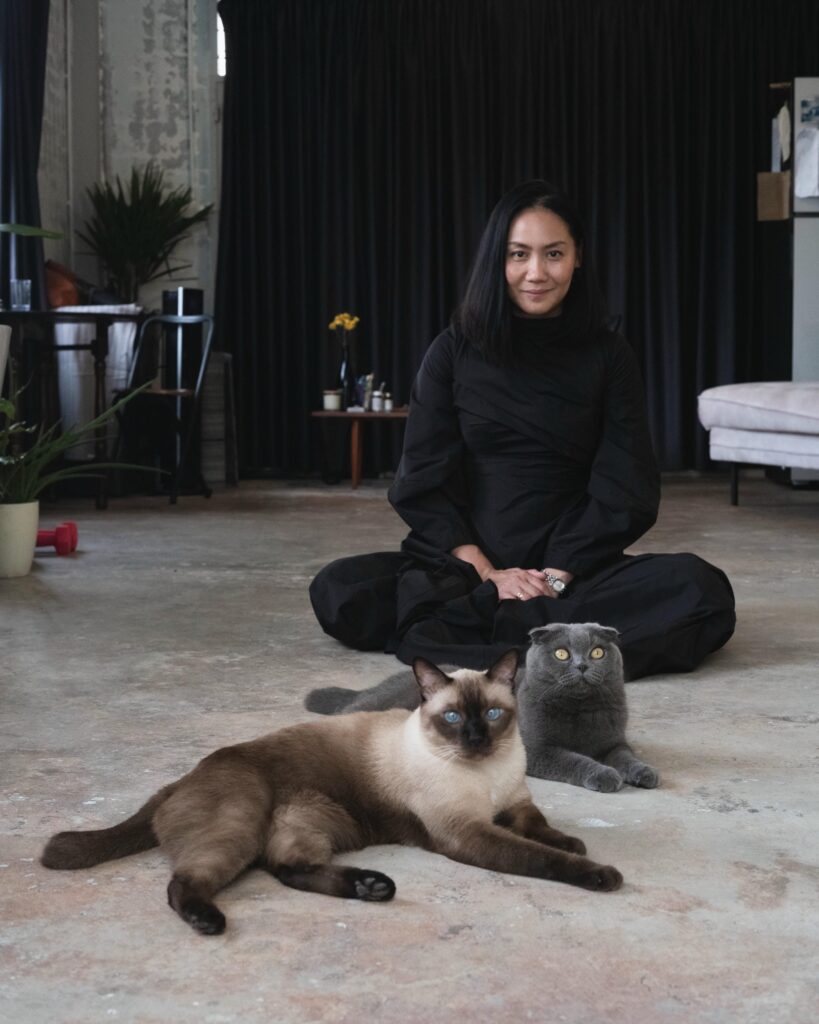 Dujdao Vadhanapakorn sitting cross-legged on the floor behind two cats.