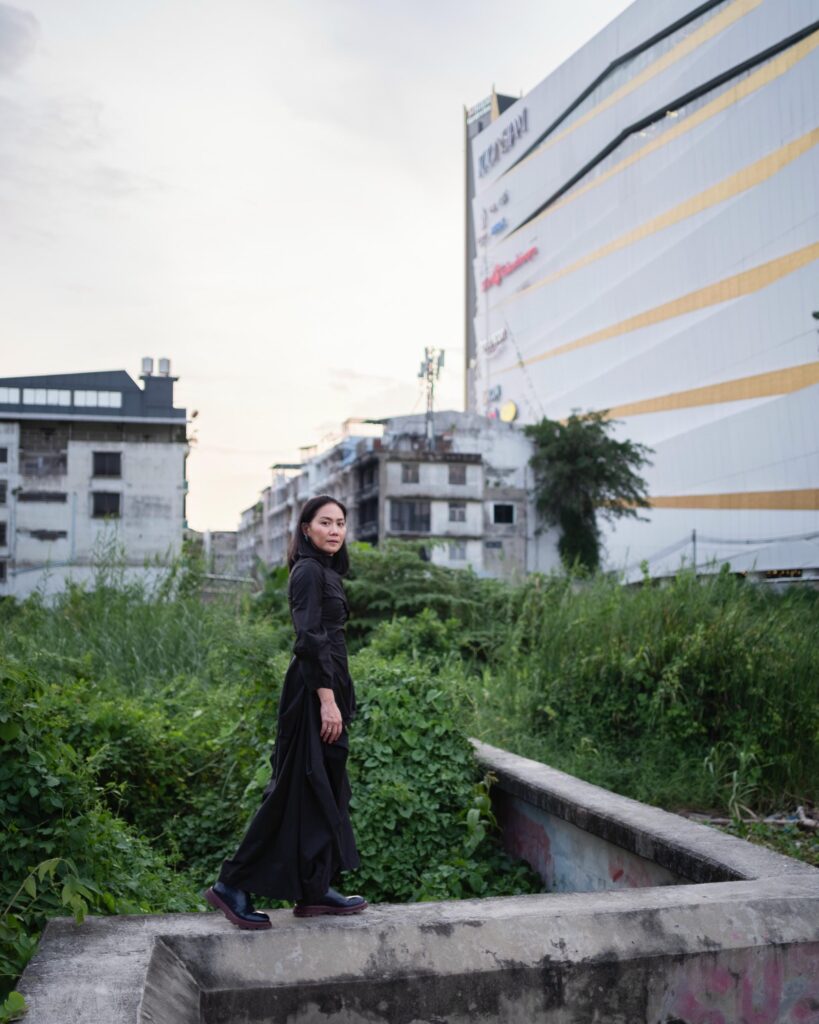 Dujdao Vadhanapakorn walking along a low concrete wall beside an overgrown Bangkok lot.