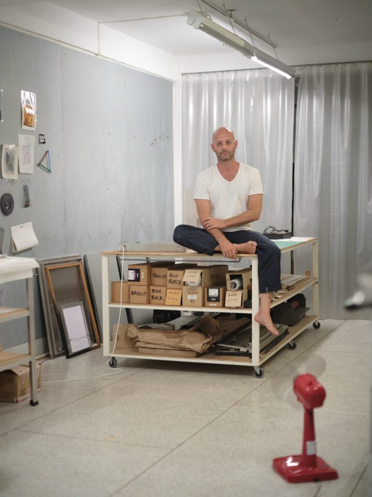 François Langella sitting on a workbench among boxes and framed works in his studio.
