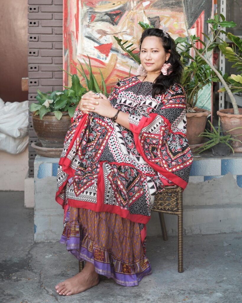 Gabriela Moriarty seated in a patterned dress beside plants and an abstract painting.