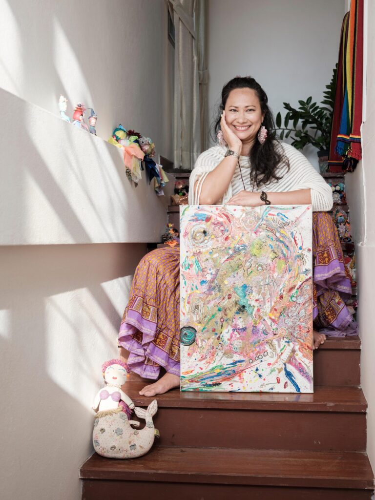 Gabriela Moriarty seated on wooden stairs with a colourful painting in front of her.