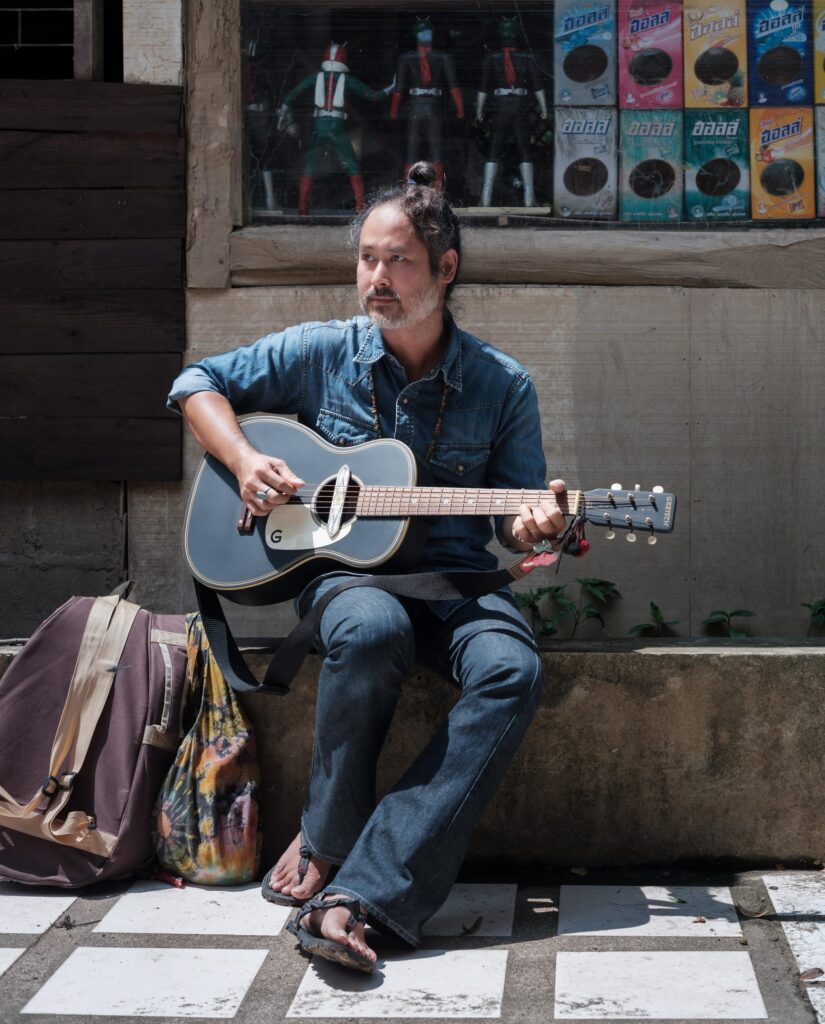 Gene Mahasmut playing an acoustic guitar while seated outside a wooden building.