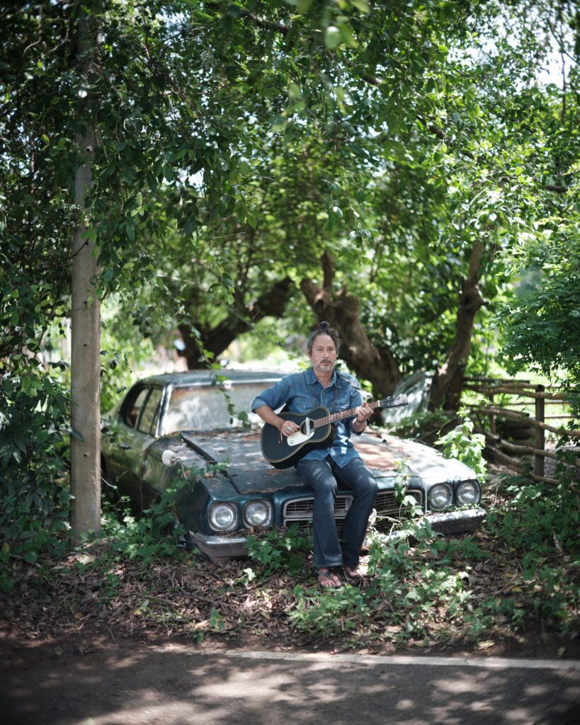 Gene Mahasmut playing guitar while seated on an old car beneath trees.