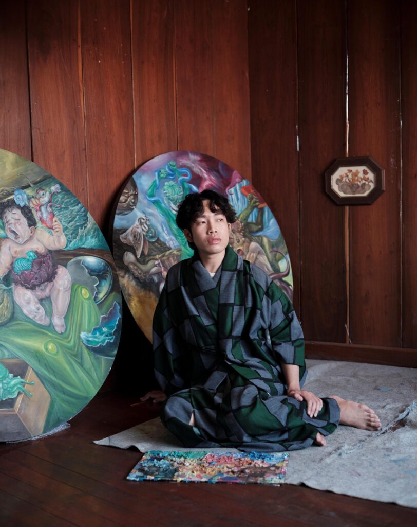 Jakkapan Mantapan sitting on the floor beside large oval paintings and a paint palette.
