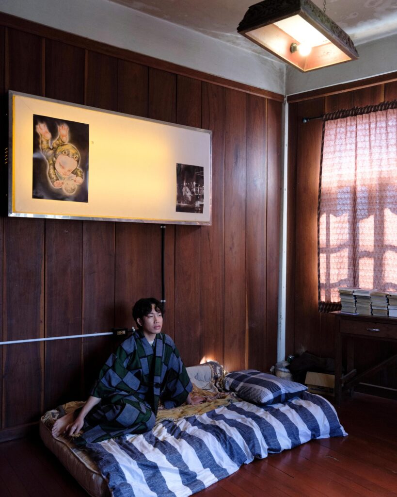Jakkapan Mantapan seated on a striped floor mattress beneath displayed artworks.