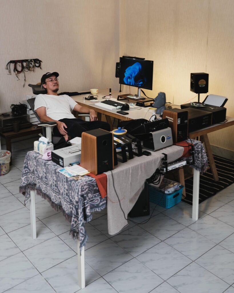 Jay Vatanakuljaras sitting at a studio desk surrounded by speakers, records and audio equipment.