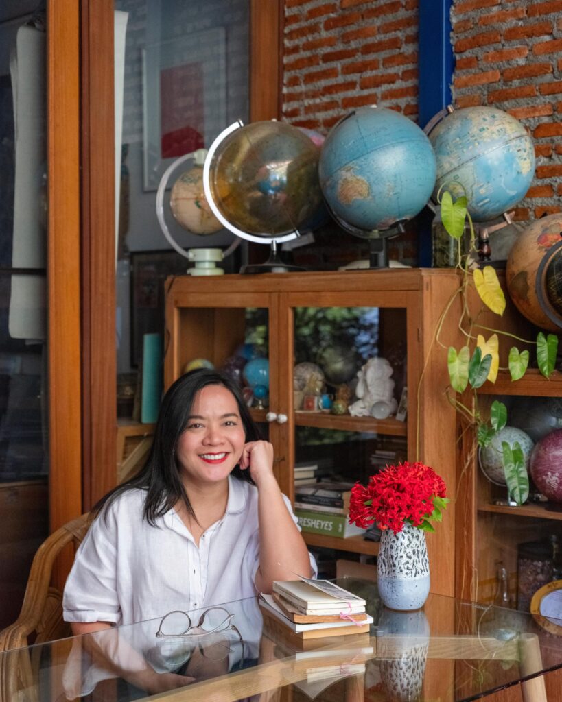 Jiratchaya Pripwai seated at a glass table beside books and red flowers, with globes behind her.
