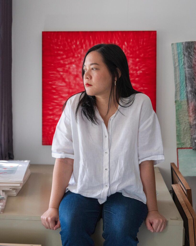 Jiratchaya Pripwai seated in her studio with a red painting on the wall behind her.