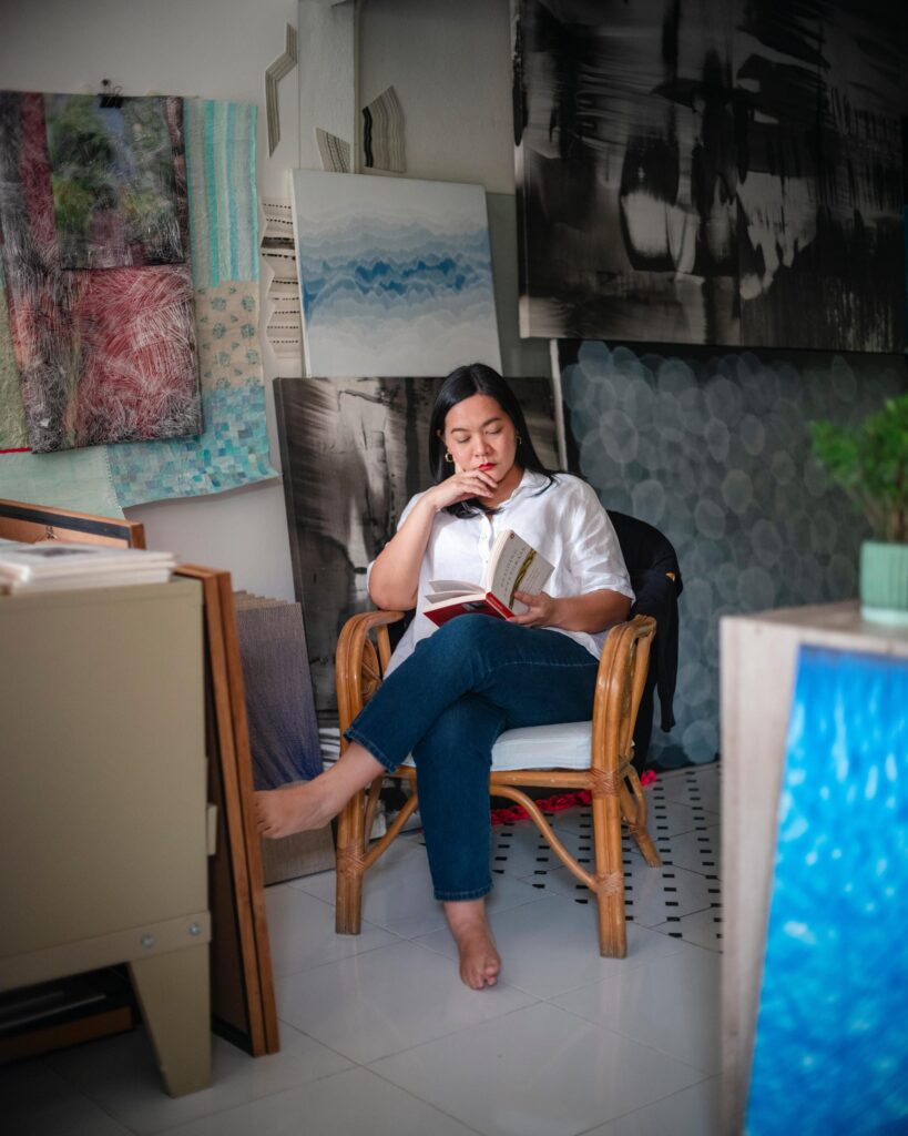 Jiratchaya Pripwai reading in a chair surrounded by paintings in her studio.
