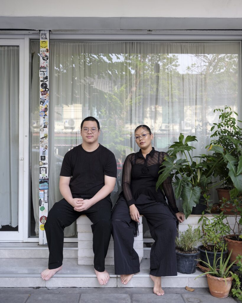 Martha and Ryan seated barefoot on the front steps of their home, with potted plants beside the doorway.