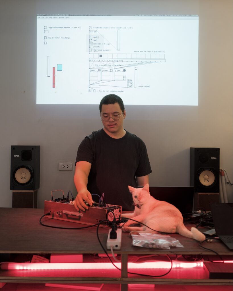 Ryan working at an electronic music desk beside a white cat, with code projected on the wall.