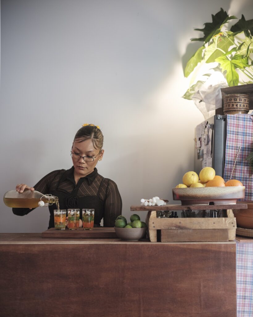Martha pouring from a glass bottle into patterned drinking glasses on a wooden bar.