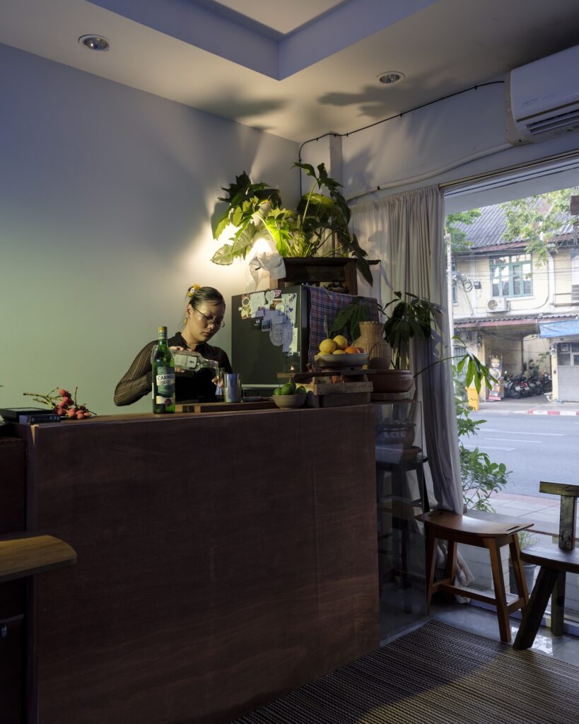 Martha pouring into a metal cup behind a wooden bar beside an open doorway.