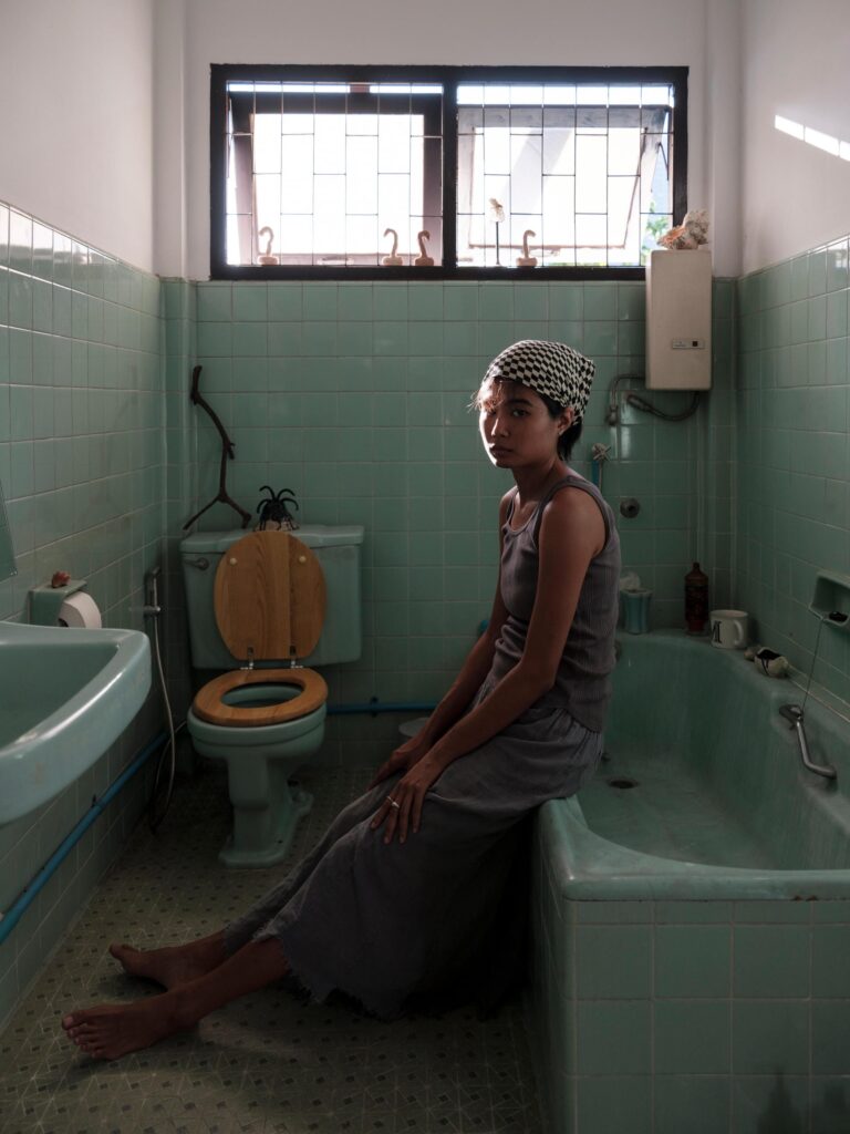 MM.Kosum seated on the edge of a bathtub in a green-tiled bathroom.