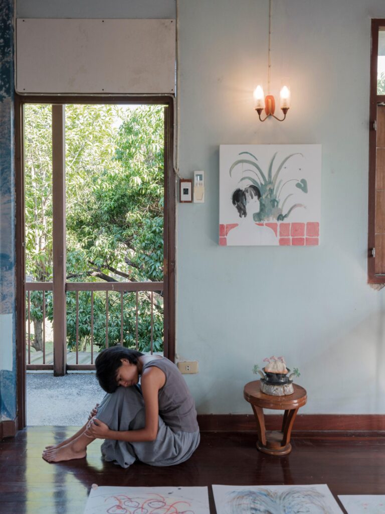 MM.Kosum sitting on a wooden floor beside an open balcony door and drawings.
