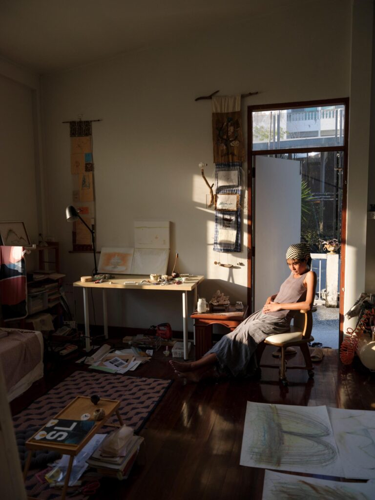 MM.Kosum seated in sunlight among drawings and materials in her Bangkok studio.