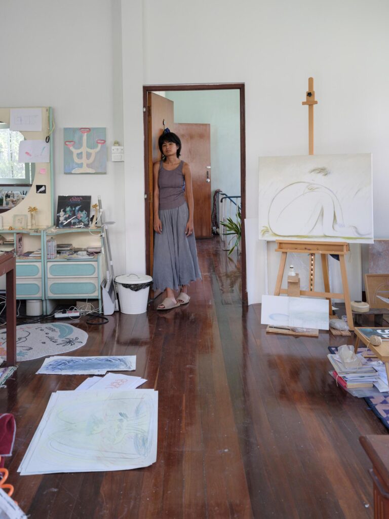 MM.Kosum standing in a studio doorway with drawings spread across the wooden floor.