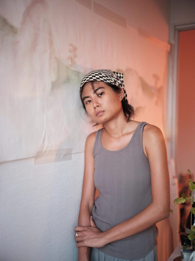 MM.Kosum wearing a patterned headscarf beside a drawing on the wall.