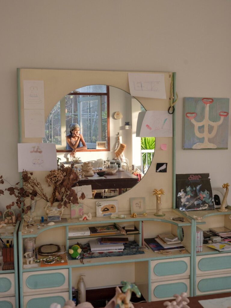 MM.Kosum reflected in a mirror above a dressing table covered with artworks and objects.