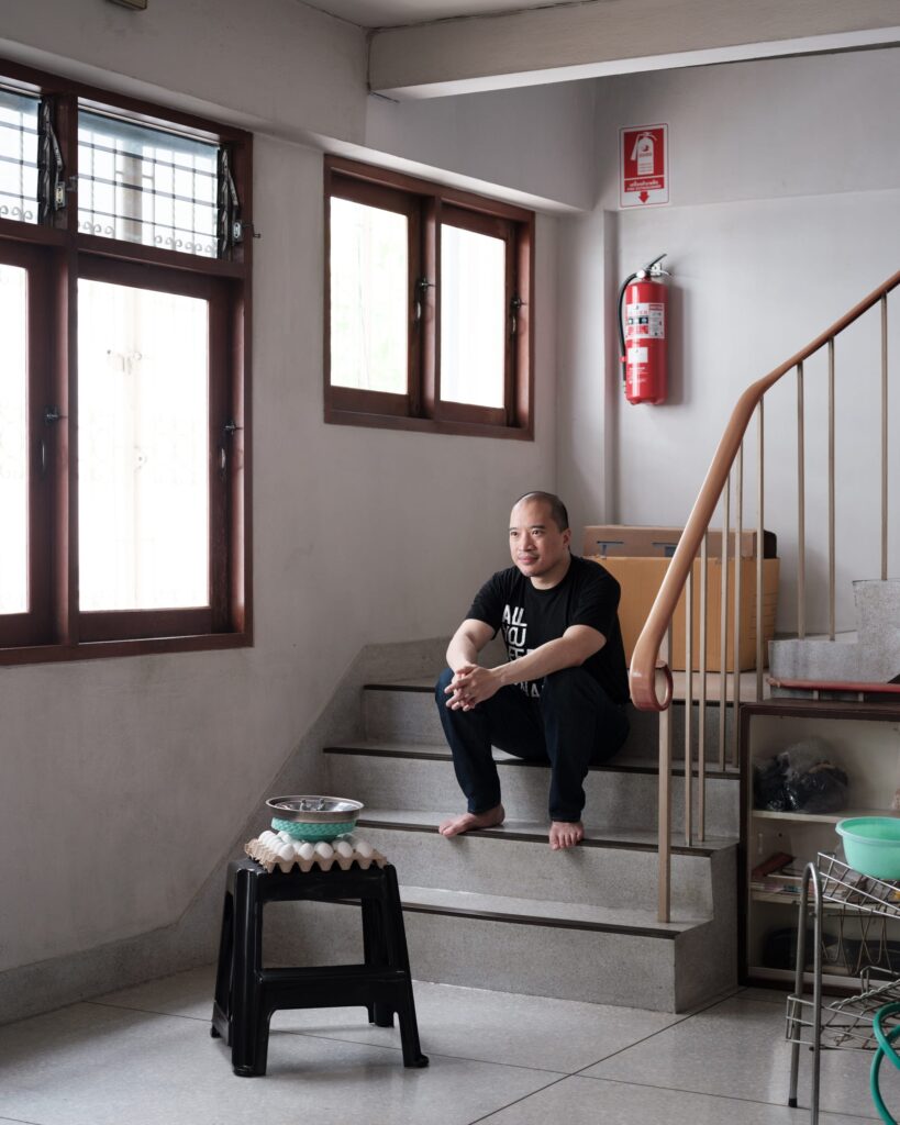 Parinot Kunakornwong sitting barefoot on stairs beside windows in his Bangkok studio.