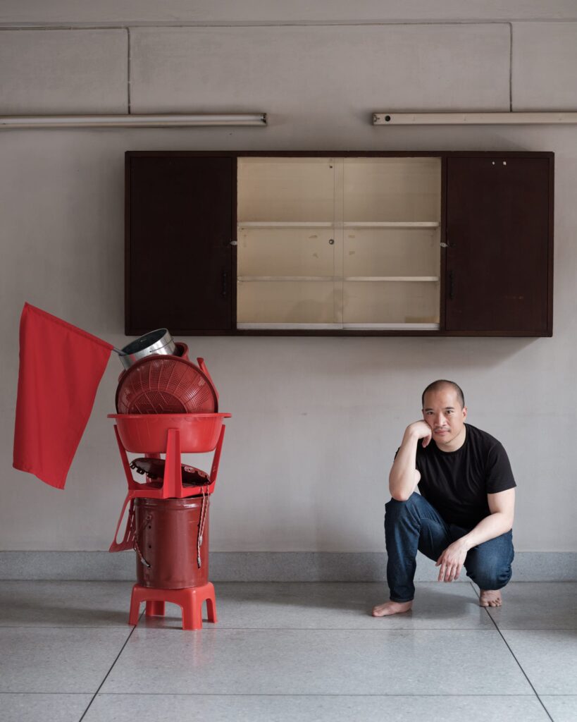 Parinot Kunakornwong crouching beside a red assemblage beneath an open wall cabinet.
