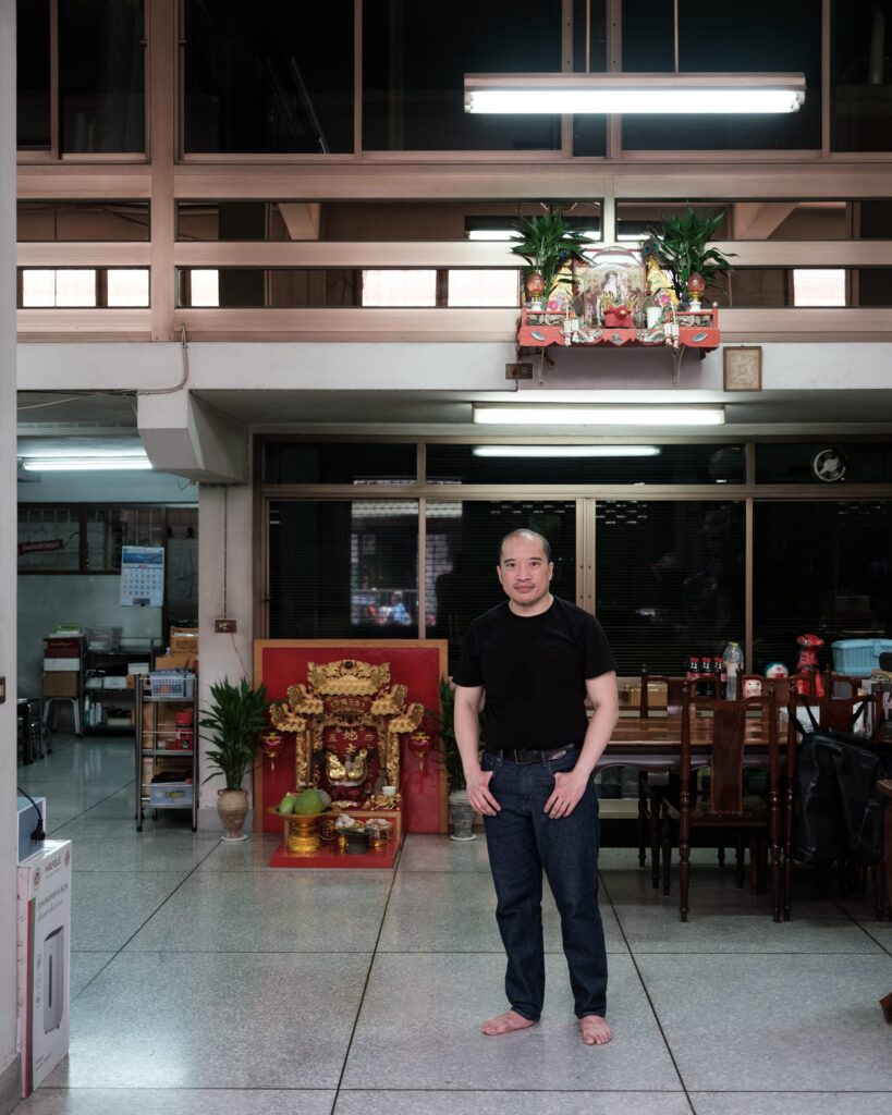 Parinot Kunakornwong standing barefoot inside his Bangkok home with a red and gold shrine behind him.