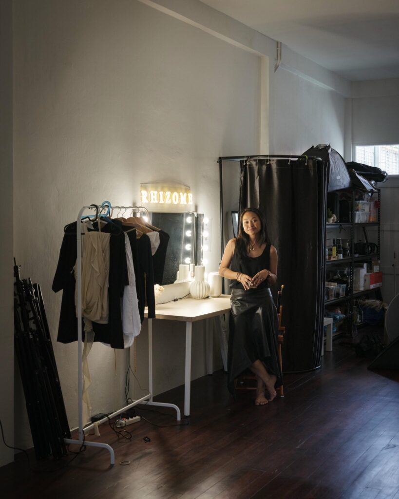 Ploy Phutpheng standing beside a lit dressing-table mirror and clothing rack.