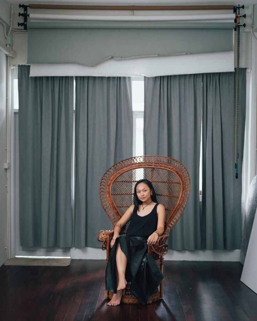 Ploy Phutpheng seated in a wicker chair beneath rolls of photographic backdrops.