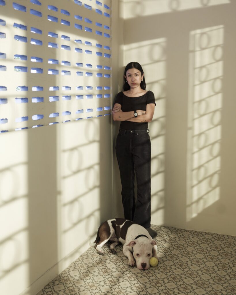 Pokchat standing in patterned sunlight with her dog lying at her feet