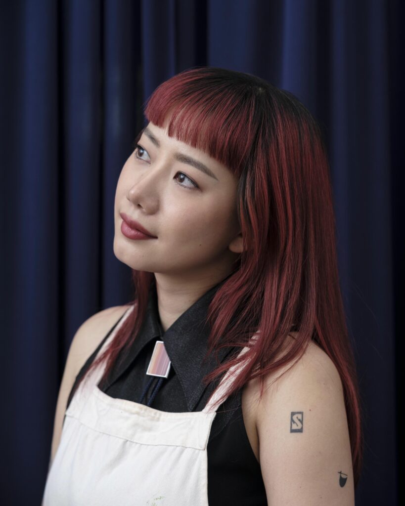 Close portrait of Sandy Chuchat in a paint-marked apron against a dark curtain.