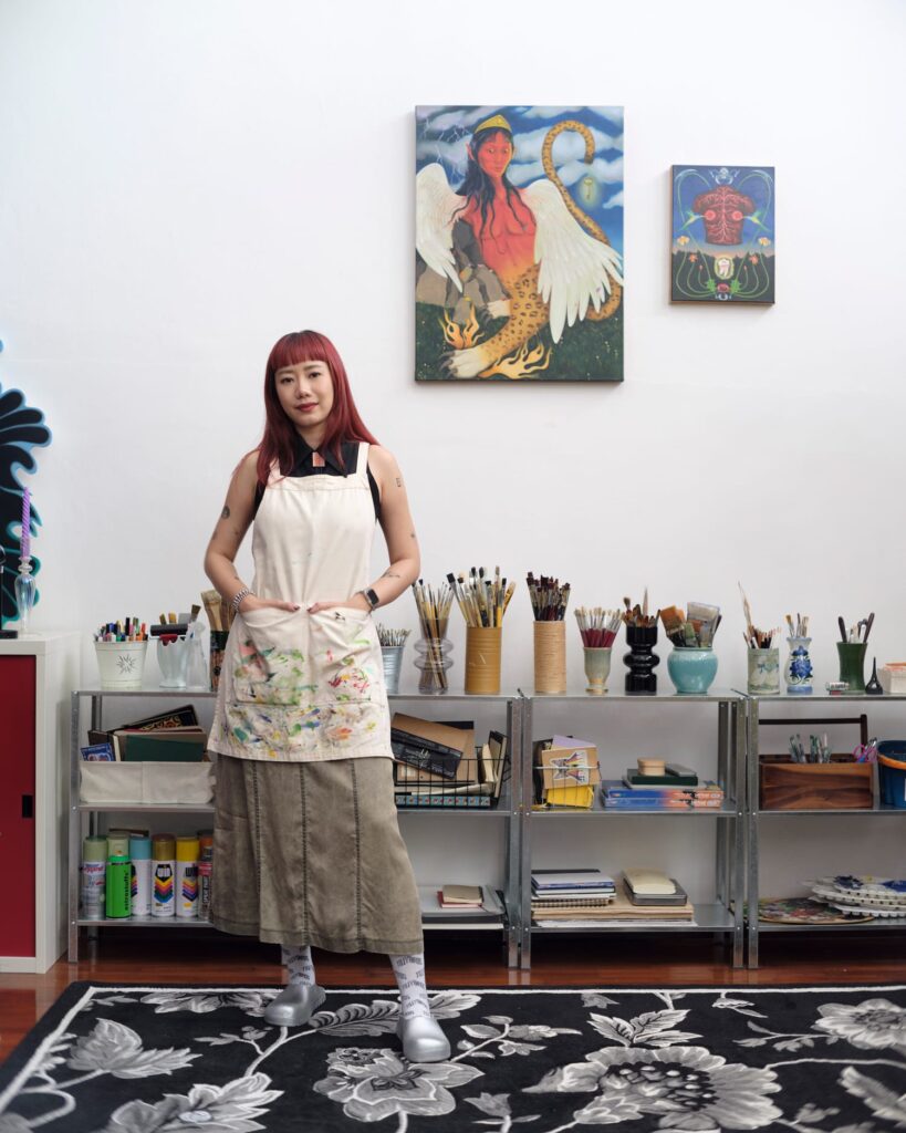 Sandy Chuchat standing beside shelves of brushes and paint beneath displayed paintings.