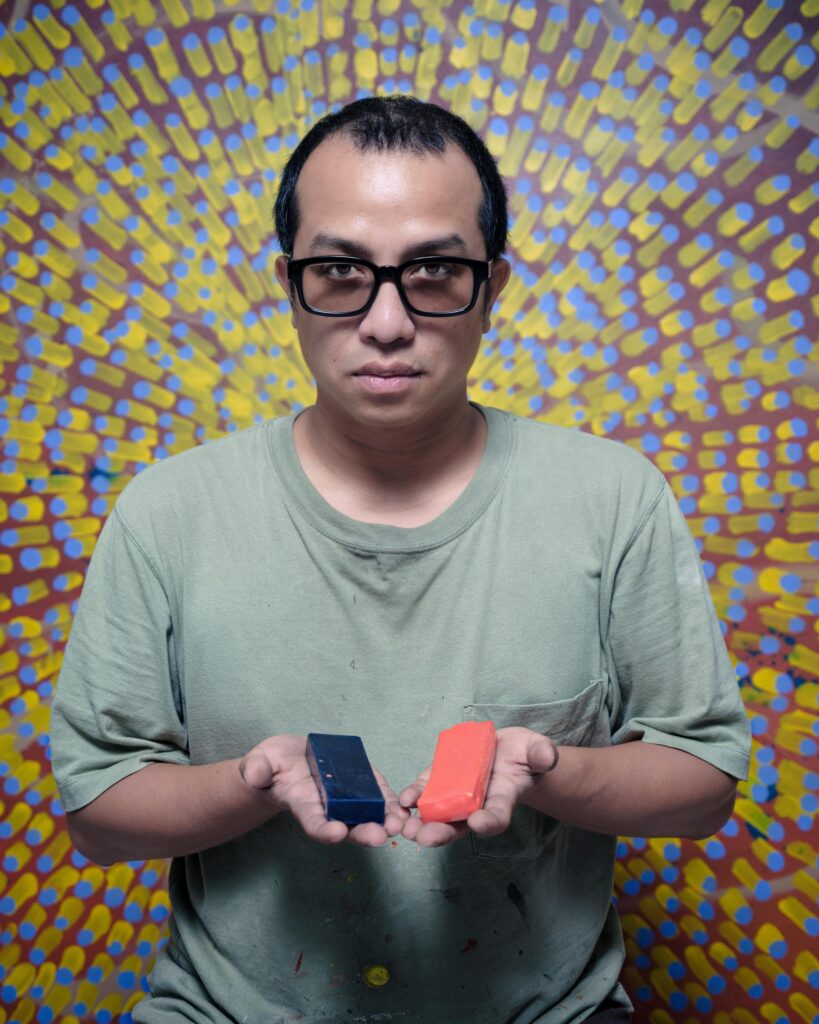 Tada Hengsapkul holding blue and red rectangular objects in front of a colourful patterned artwork.