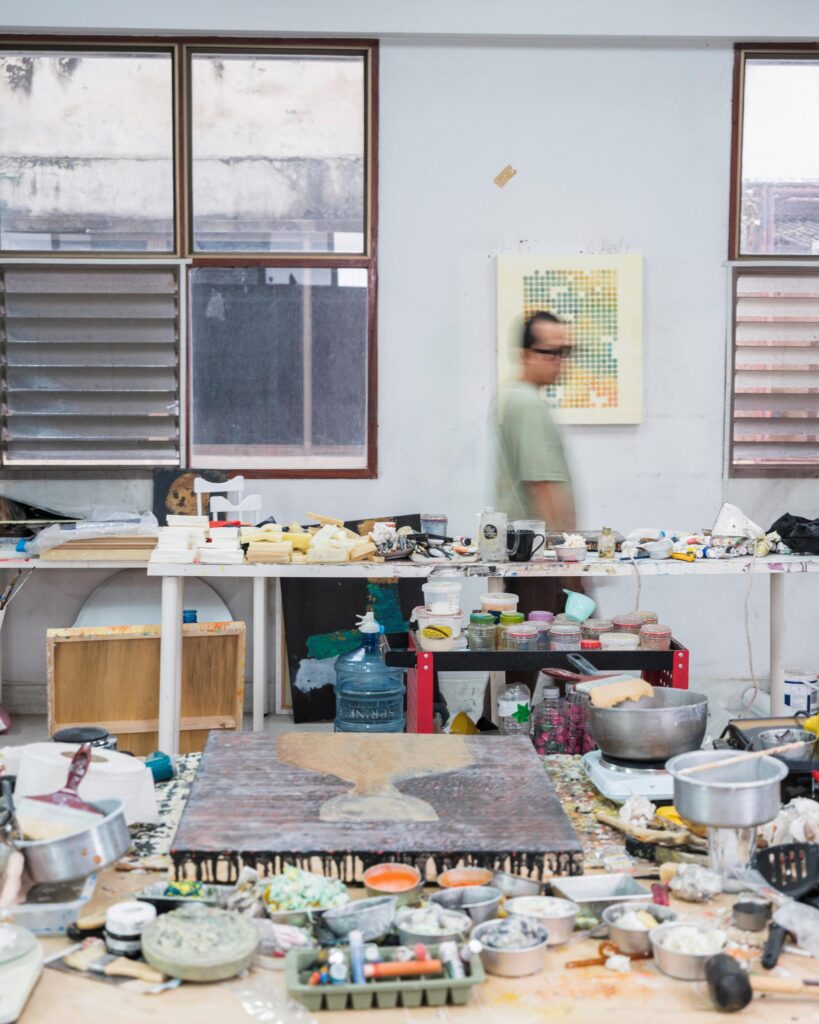 Tada Hengsapkul moving behind a studio worktable covered with bowls and painting materials.